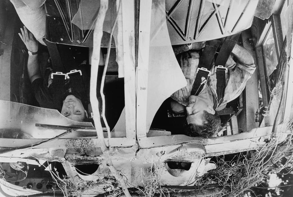 Black-and-white screen grab from Air America showing two pilots hanging upside down in the wreckage of a crashed aircraft, still strapped into their seats amid twisted metal, broken panels, exposed wiring, and debris.